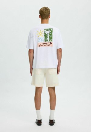Homme portant un T-shirt blanc avec un motif coloré sur le thème de la plage dans le dos, un short crème, des chaussettes blanches et des chaussures noires, debout de dos.