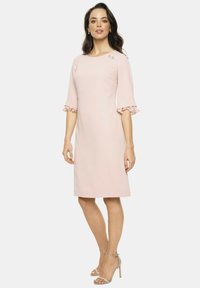 Jasnoróżowa sukienka z dekoltem w serek, rękawami do trzech czwartej długości i falbaniastymi mankietami. Posiada dekoracyjne akcenty na ramieniu oraz smukłą sylwetkę.