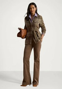 Γυναίκα σε καφέ καρό μπερδεμένο σακάκι με ζώνη και καφέ παντελόνι, κρατώντας μια ταμπά τσάντα, στέκεται σε έναν απλό ανοιχτό φόντο.