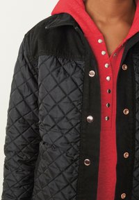 Chaqueta negra acolchada con hombros de pana, broches en color oro rosa y una sudadera roja debajo con una tapeta abotonada y textura suave.