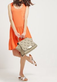 Oranžové bezramínkové šaty s kapsou, s vrstvenou sukní. Béžová kabelka s embosovanými vzory a zlatým zapínáním, doplněná o bílé sandály.