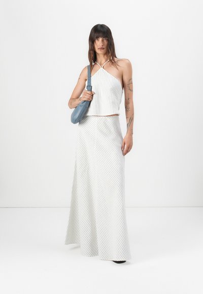 Baltas un krēms svītrainas halter stila cropped krekls, kas apvienots ar atbilstošu garu svārku un gaismas zila rokassomu. Izvietots uz vienkārša fona.