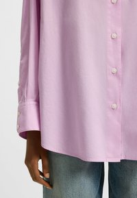 Camicia oversize rosa chiaro con chiusura a bottoni e dettagli in perle sui polsini, abbinata a denim azzurro.