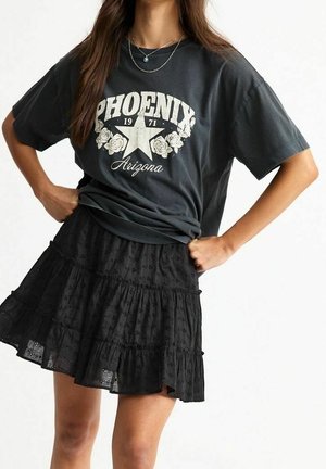 Kobieta ubrana w czarny t-shirt z napisem "Phoenix Arizona" i czarną, warstwową spódnicę, pozująca z rękami na biodrach na jednolitym tle.