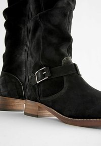 Bottes noires en daim hautes jusqu'au genou avec un motif matelassé au talon, dotées d'une fermeture éclair sur le côté et d'une boucle en métal argenté sur un talon en bois.