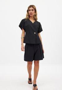 Melna divdaļīgā apģērba komplekts ar V-veida kakla izgriezumu, īsām piedurknēm un plīša šortiem, ar dekoratīvu pogu detaļu un gludu tekstūru.