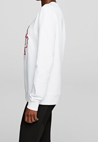 Sudadera blanca de manga larga, con cuello redondo, diseño bordado en rojo y azul, textura suave y puños acanalados, combinada con leggings negros.