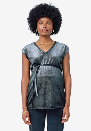 Frau mit kurzen lockigen Haaren, die ein ärmelloses, metallic-graues Wickeltop trägt, das an der Taille gebunden ist, und dunkle Hosen, stehend vor weißem Hintergrund.