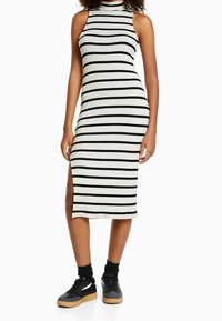 Robe midi à rayures noir et blanc, en tissu côtelé, avec un col haut, une fente sur le côté et une coupe ajustée. Assortie à des baskets noires et des chaussettes mi-mollet.