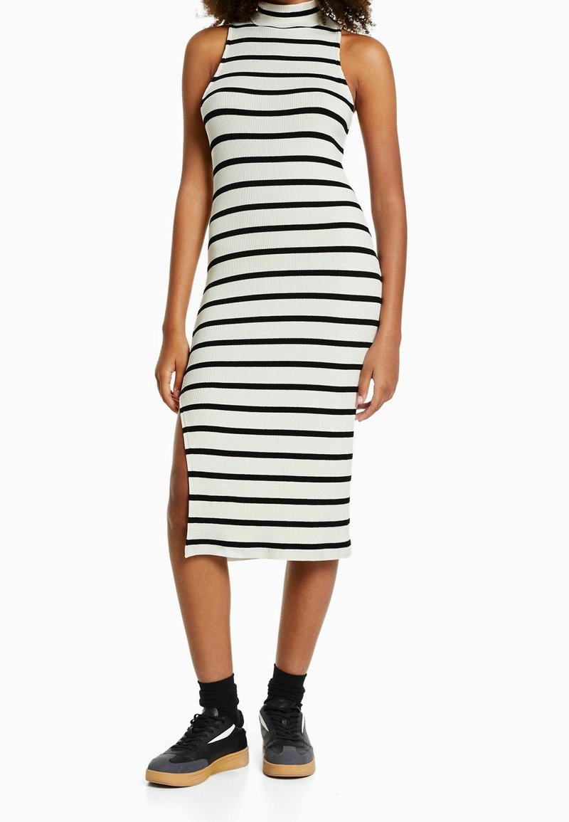 Robe midi à rayures noir et blanc, en tissu côtelé, avec un col haut, une fente sur le côté et une coupe ajustée. Assortie à des baskets noires et des chaussettes mi-mollet.