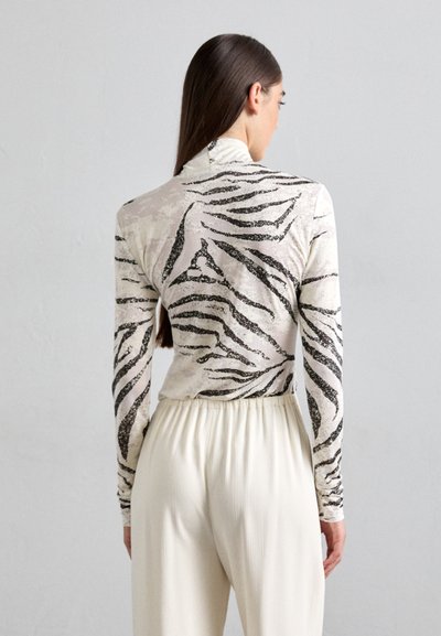 Dugmad s dugim rukavima koje se ističe prozirnom bijelom tkaninom s crnim prugama u zebrinom uzorku. Upareno sa svijetlim, plisiranim hlačama.
