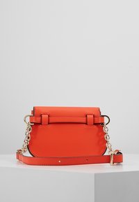 Sac à main en similicuir orange avec une forme structurée, des éléments en chaîne et en sangle, un design rectangulaire et une fermeture par bouton-pression à l'avant.