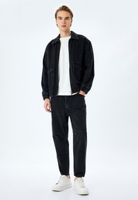 Chaqueta de mezclilla negra con puños elásticos, llevada sobre una camiseta blanca, combinada con pantalones vaqueros negros y zapatillas blancas. El modelo está de pie contra un fondo claro.