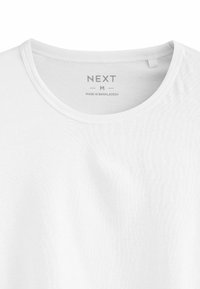 Fehér, pamut póló kerek nyakkivágással, minimális textúrával, rajta a "NEXT M" és a "MADE IN BANGLADESH" felirattal.