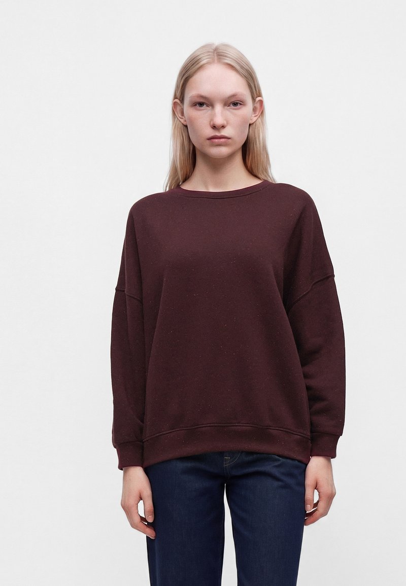 Femme aux cheveux blonds raides porte un sweat-shirt marron foncé ample et un jean bleu foncé, debout devant un fond clair uni.