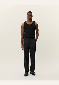 Black tank top paired with high-waisted gray trousers. The model stands in black shoes against a neutral background.