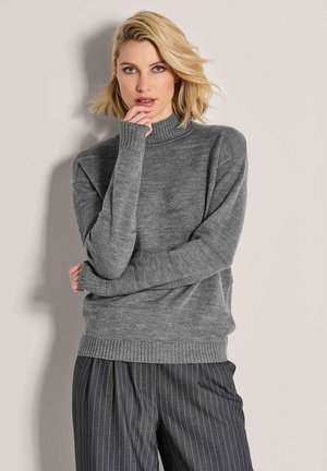 Blonde Frau mit schulterlangem Haar trägt grauen Strickpullover und gestreifte Hose, posiert mit der Hand nahe dem Mund vor einem neutralen Hintergrund.