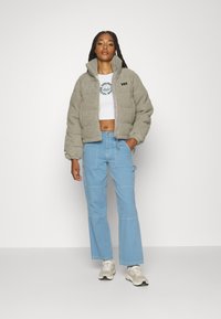 Giacca cropped grigia chiara con una texture morbida e colletto alto, abbinata a pantaloni cargo blu chiaro. T-shirt bianca con grafica sotto.
