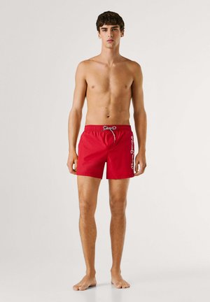 Giovane uomo in piedi a piedi nudi, indossa pantaloncini da bagno rossi con logo bianco sul lato destro, sfondo chiaro uniforme.