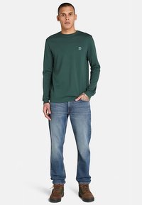 Zielona koszula z długim rękawem, okrągłym dekoltem i małym logo, zestawiona z jasnoniebieskimi dżinsami i brązowymi butami trekkingowymi. Luźny fason.