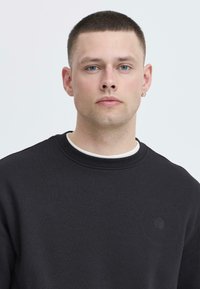 Schwarzer Sweater mit Rundhalsausschnitt und gerippten Bündchen, ausgestattet mit einem kleinen geprägten Logo auf der Brust und einer strukturierten Stoffoberfläche.