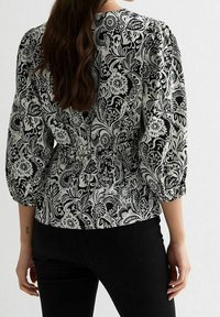 Blouse imprimée florale en noir et blanc avec manches bouffantes et taille froncée. Tissu lisse avec des motifs complexes ; associée à un pantalon noir.