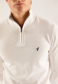 Pullover bianco lavorato a maglia con collo a zip quarti, che presenta un ricamo di un piccolo uccello blu sul petto e polsini a costine.