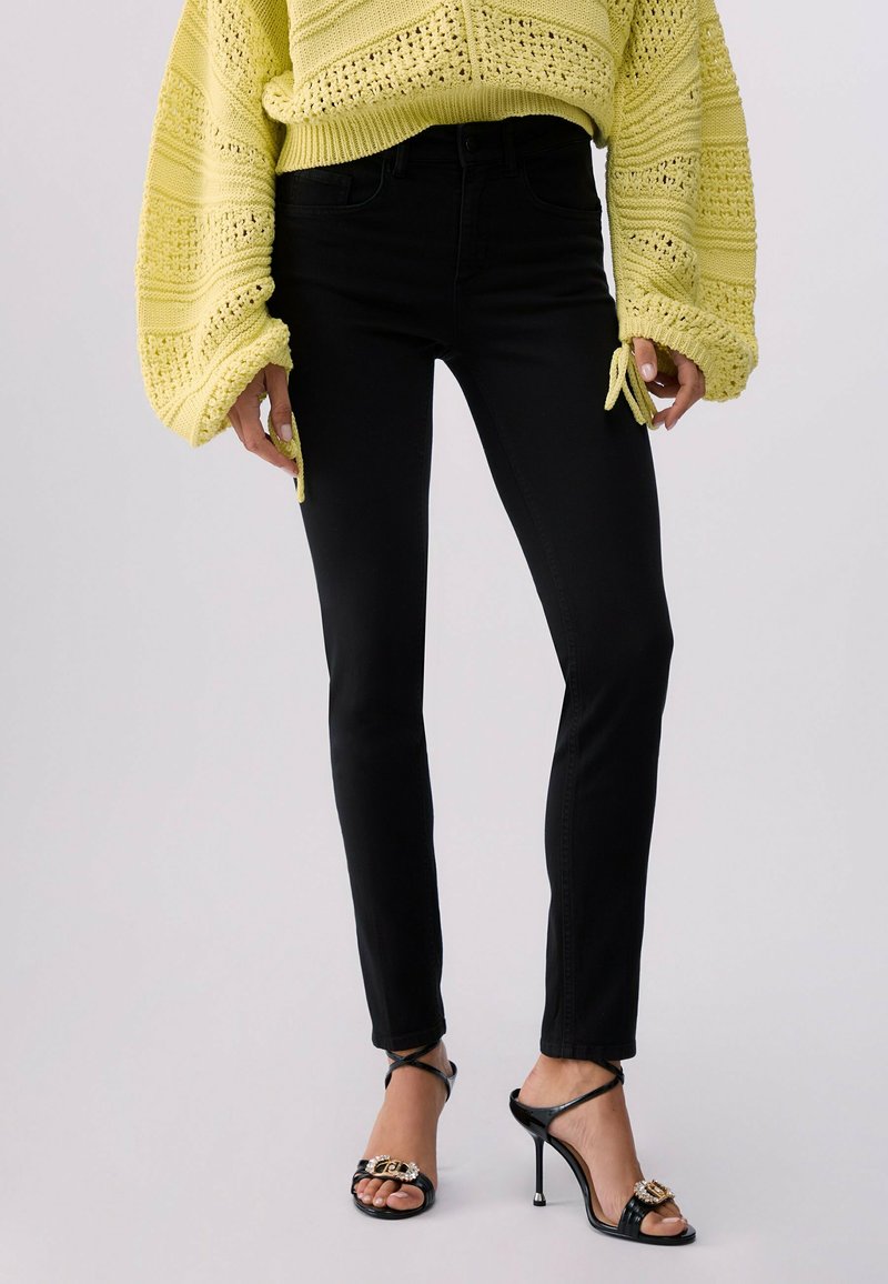 Femme portant un jean skinny noir, des sandales à talons hauts noires avec des ornements, et un pull en crochet jaune à manches larges.