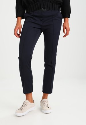 Jalat, joissa on slim fit -tyyliset tummansiniset housut ja beiget nauhalliset tennarit, yhdistetty mustaan toppiin, seisovat valkoista taustaa vasten.