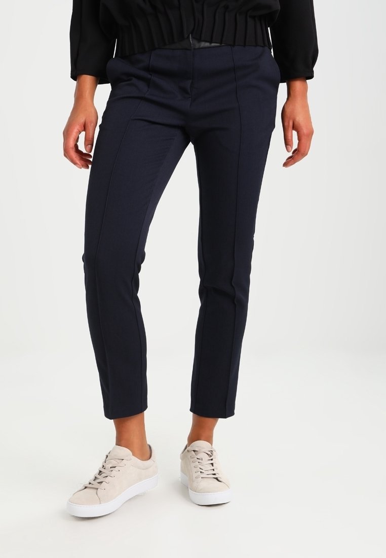 Jambes portant un pantalon bleu marine slim et des baskets beiges à lacets, associées à un haut noir, debout sur un fond blanc.