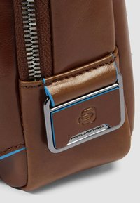 Hoek van een bruine leren tas met een zilveren ritssluiting en plaat, met blauwe accenten. Soepele textuur met een gestructureerd ontwerp.