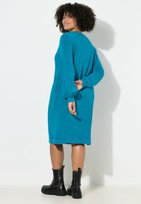 Kobieta z kręconymi włosami, ubrana w turkusową sukienkę z długim rękawem i czarne, ćwiekowane botki za kostkę, stoi z jedną ręką za plecami.