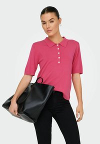 Donna che indossa una polo rosa a maniche corte e pantaloni neri, mentre tiene una grande borsa a tracolla in pelle nera su uno sfondo neutro.