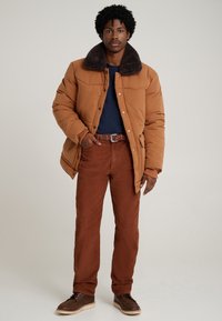 Ruda pūsta striukė su tamsiu kailio apykakle, derinama su rūdžių spalvos korduro pėdkelnėmis. Modelis vilki tamsiai mėlyną marškinius ir rudus odinius batus.