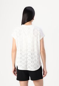 Blouse blanche à manches courtes en tissu brodé, avec un col rond et une coupe décontractée, portée sur un short noir.