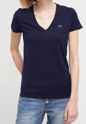 Kobieta w granatowym T-shircie z krótkim rękawem i dekoltem w serek z małym logo na piersi oraz jasnoniebieskich dżinsach, stojąca na jednolitym tle.