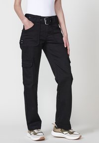 Persona que lleva pantalones cargo negros con cinturón y bolsillos, combinados con zapatillas blancas y verde oliva, de pie contra un fondo liso.