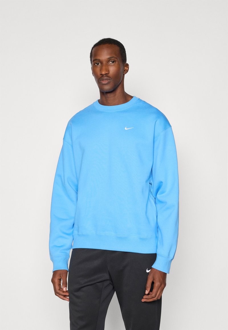 Jasnoniebieska bluza z okrągłym dekoltem i ściągaczami, z małym białym logo Nike na piersi. Ciemne spodnie poniżej.