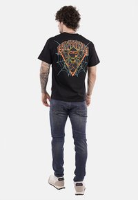 T-shirt in cotone nero con una grafica colorata di un ragno e una ragnatela sul retro, con una vestibilità comoda e maniche corte, abbinata a jeans scuri.