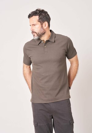 Man met baard die een bruin gestructureerd poloshirt en donkere cargobroek draagt, staat met zijn handen achter zijn rug, kijkend naar links.