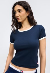 Γυναίκα με σγουρά σκούρα μαλλιά που φοράει μπλε ναυτικό κοντομάνικο τοπ με λευκή διακόσμηση και μπλε ναυτικό παντελόνι που ταιριάζει.