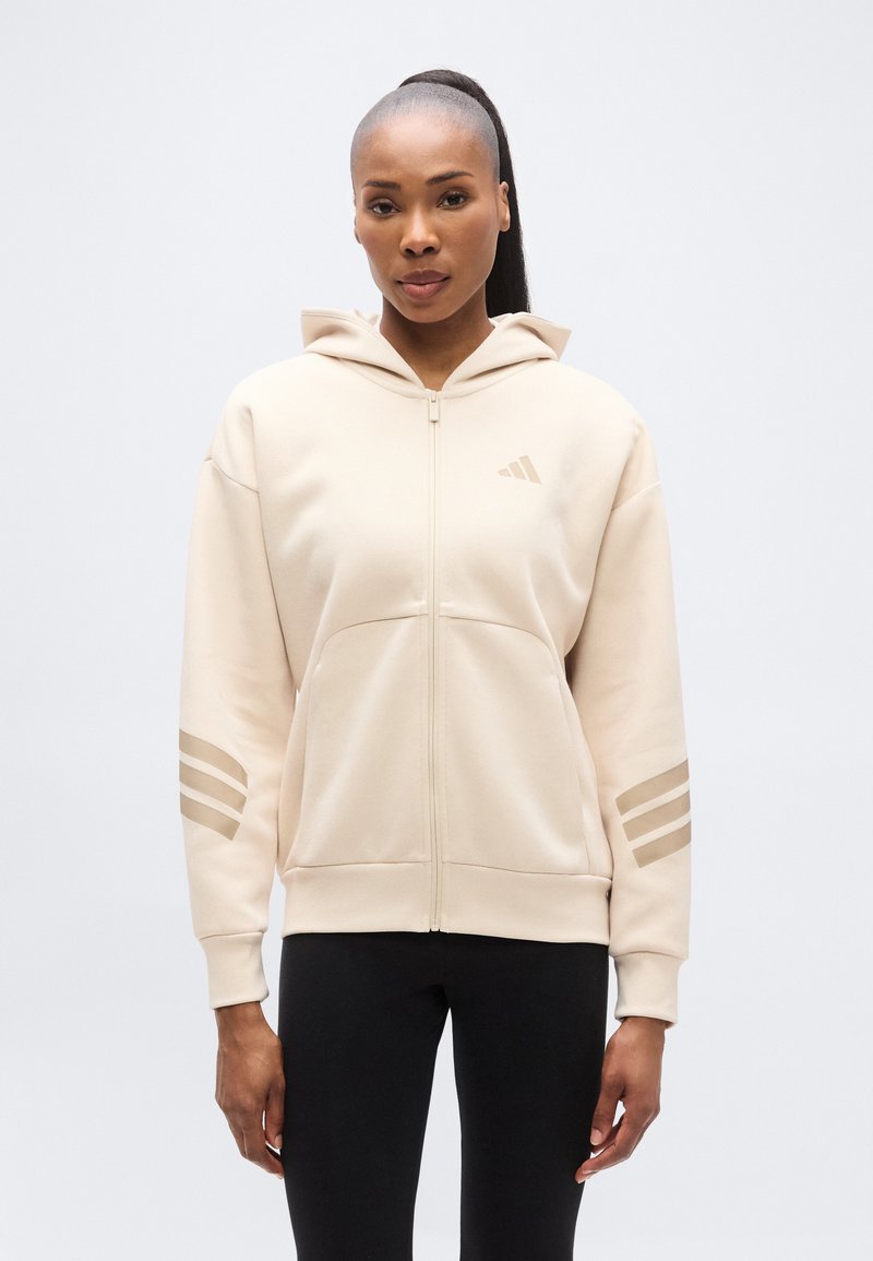 Μπεζ φούτερ με φερμουάρ και κουκούλα, κατασκευασμένο από απαλό ύφασμα. Διαθέτει διακριτικό σχέδιο με τρεις ρίγες στα μανίκια και μικρό λογότυπο στο στήθος.