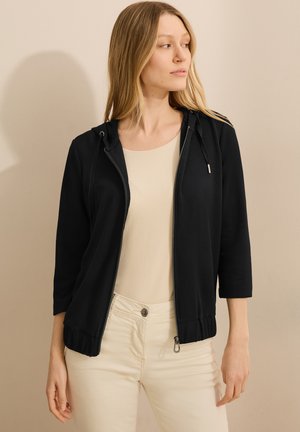 Black zip-up hoodie with a hood, three-quarter sleeves, and elastic band at the hem. Worn over a beige top, featuring smooth fabric texture.