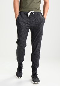Men's black jogger pants with a textured finish, elastic waistband, white drawstring, and tapered legs, paired with black athletic shoes.