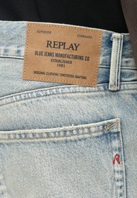 Jeans de ganga azul claro com uma etiqueta de couro rotulada "REPLAY", com detalhes de costura e um pequeno "R" vermelho no bolso.