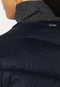 Žena oblečená v námořnickém péřovém kabátu s třpytivým černým vysokým límcem a zlatým kulatým náušnicí, z pohledu zezadu a z boku.