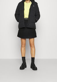 Czarna puchowa kurtka, żółty sweter, czarna spódnica i czarne sznurowane botki. Posiada teksturowany materiał, zamek błyskawiczny oraz akcenty z logo.