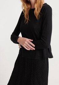 Musta pitkähihaisvaate, joka on valmistettu pehmeästä kankaasta; siinä on liehukassa ja pyöreä pääntie, yhdistetty mustan hameen kanssa, jossa on hienovaraista pintarakennetta.