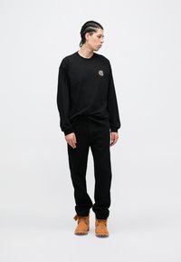 Schwarzes Langarmshirt mit kleinem Logo, kombiniert mit schwarzer Hose und beige Stiefeln. Das Model steht seitlich vor einem schlichten weißen Hintergrund.