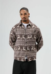 Chemise en flanelle marron avec des motifs de rennes blancs et des motifs géométriques, dotée d'un col, d'une fermeture boutonnée et d'une poche poitrine unique.